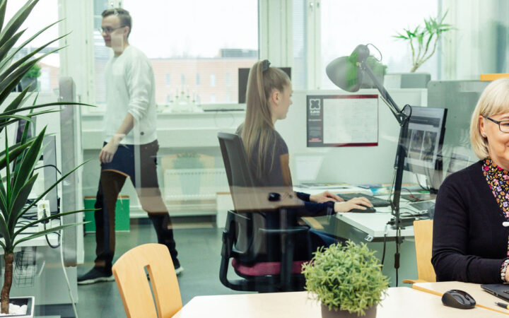 Kliininen asiantuntija YAMK -opiskelija työskentelee koneella toimistossa, jonka lasiseinän takana näkyy muita opiskelijoita työssä