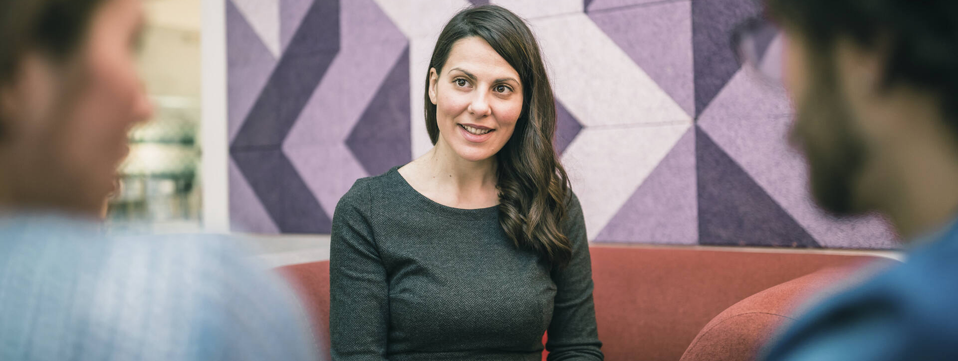 Nuori nainen istuu sohvalla ja vastapäätä istuu selin kaksi miestä.