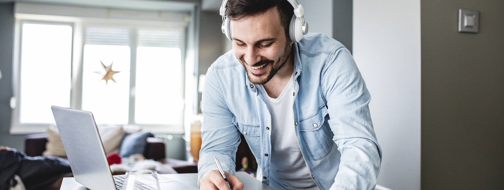 Mies kirjoittaa kuulokkeet korvissaan paperille, edessä on tietokone pöydällä.