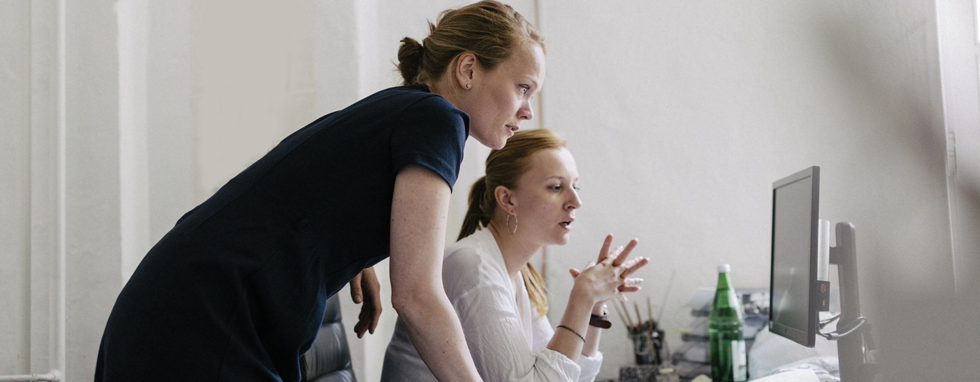 Kaksi naista tietokoneen äärellä. Toinen istuu työtuolissa, toinen seisoo vieressä nojaten toisella kädellä työpöytään.