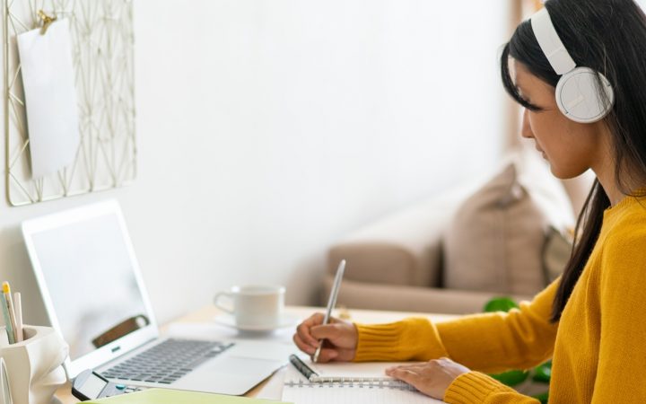 Nainen opiskelemassa kotonaan kannettavan tietokoneen äärellä. Naisella on kuulokkeet korvillaan ja hän tekee muistiinpanoja.