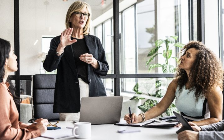 Työkollegat neuvotteluhuoneessa. Muut istuvat, yksi seisoo pöydän päässä johtaen tapeamista.