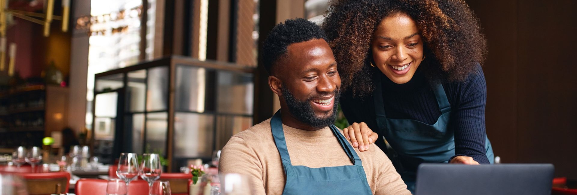 Kaksi hymyilevää henkilöä työskentelemässä kannettavalla tietokoneella ravintolaympäristössä pöydän ääressä. Toinen henkilö istuu edessä, toinen seisoo hänen takanaan toinen käsi hänen olkapäällään.