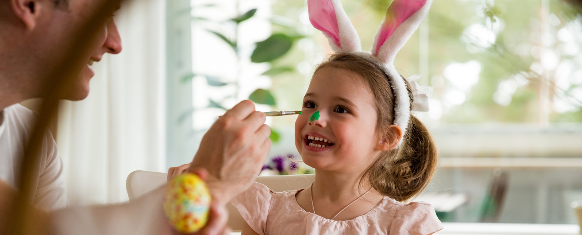 Isä ja tytär maalaamassa pääsiäismunia, ja tytön nenää vihreäksi. Tytöllä on päässään pupunkorvat,