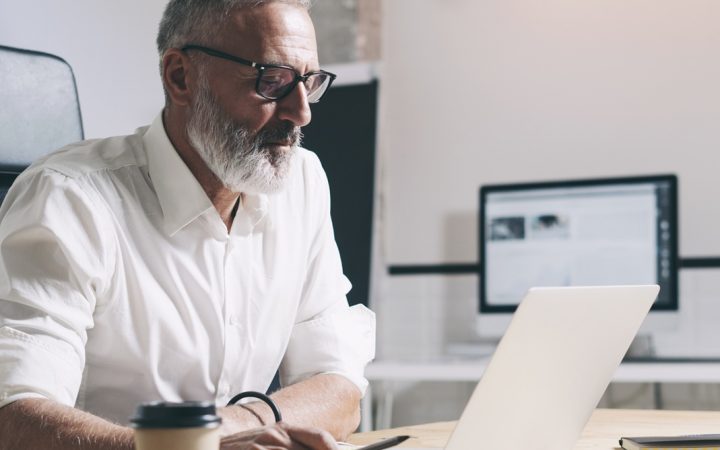 Keski-ikäinen liikemies työskentelee kannettavalla tietokoneella ja istuu puupöydän ääressä modernissa toimistotilassa.