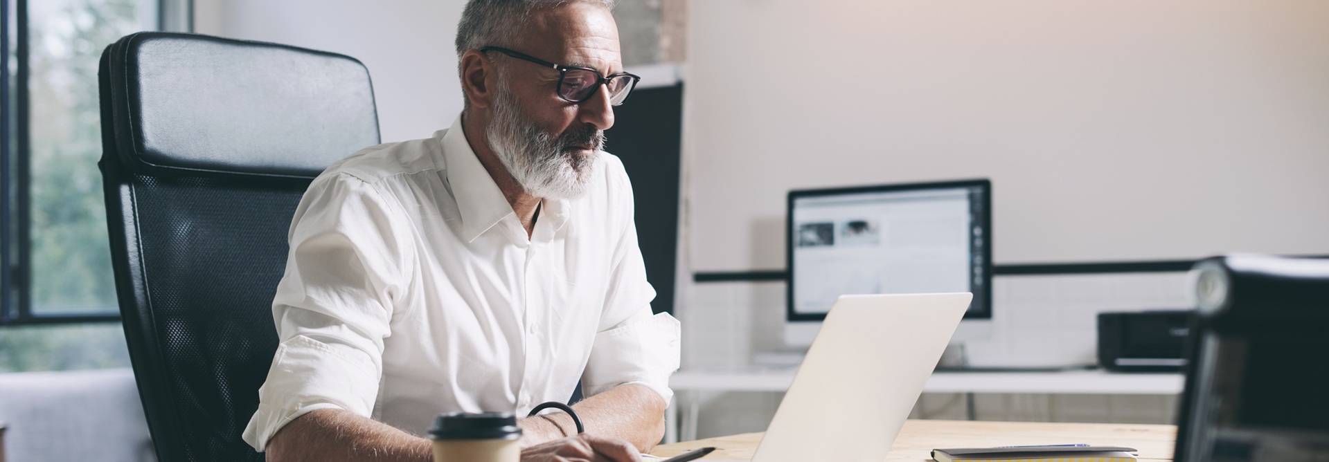 Keski-ikäinen liikemies työskentelee kannettavalla tietokoneella ja istuu puupöydän ääressä modernissa toimistotilassa.