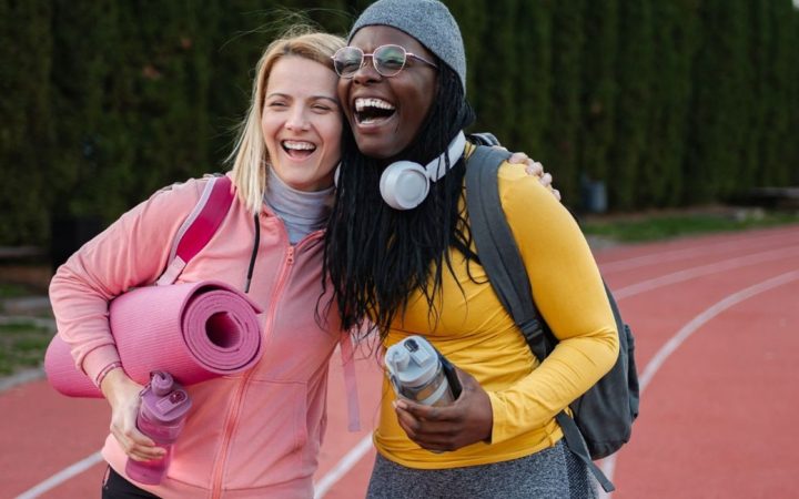 Kaksi naista urheilukentän juoksuradalla halaavat. toisella kainalossa jumppamatto, molemmilla reput ja kädessä juomapullo.
