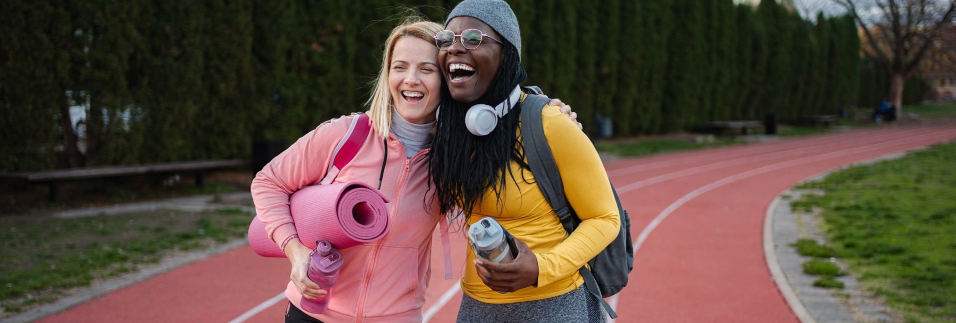 Kaksi naista urheilukentän juoksuradalla halaavat. toisella kainalossa jumppamatto, molemmilla reput ja kädessä juomapullo.