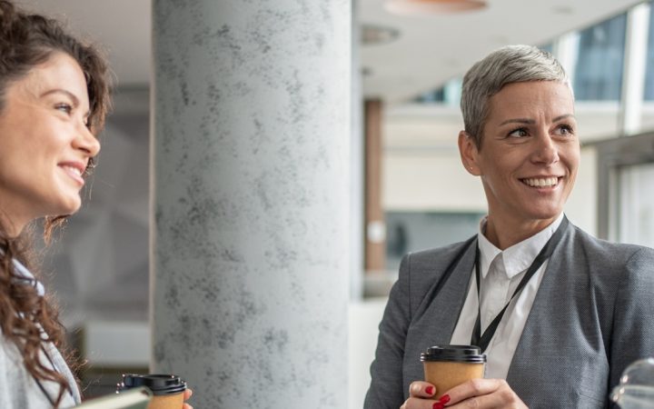 Kolme hymyilevää naista seisovat modernissa toimistotilassa kahvit kädessään.