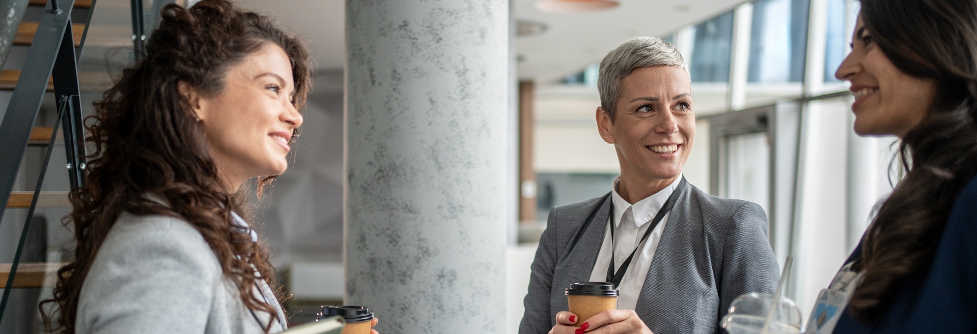 Kolme hymyilevää naista seisovat modernissa toimistotilassa kahvit kädessään.