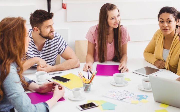 Viisi henkilöä istuu pöydän ääressä edessään laptop ja tabletti sekä posti it -lappuja.