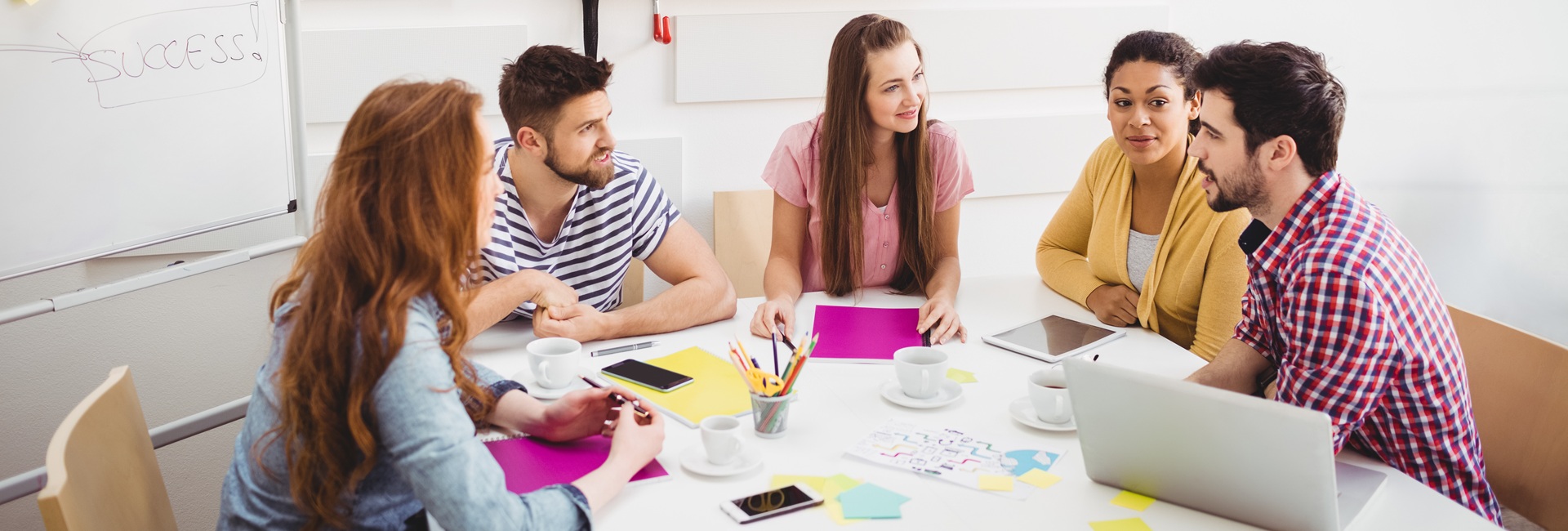 Viisi henkilöä istuu pöydän ääressä edessään laptop ja tabletti sekä posti it -lappuja.