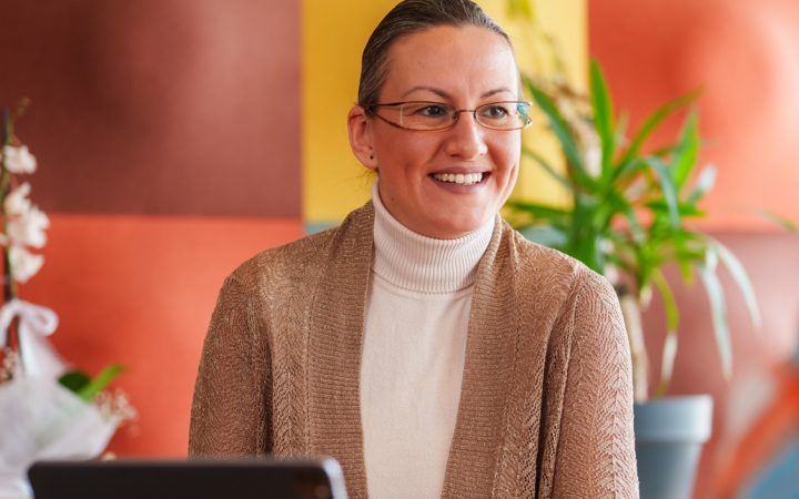 Hymyilevä silmälasipäinen nainen katsoo vastapäätä istuvaa henkilöä. Välissä pöydällä kannettava tietokone.