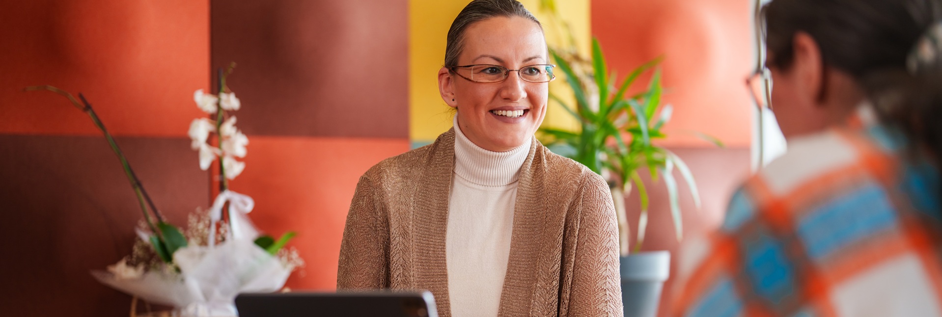 Hymyilevä silmälasipäinen nainen katsoo vastapäätä istuvaa henkilöä. Välissä pöydällä kannettava tietokone.