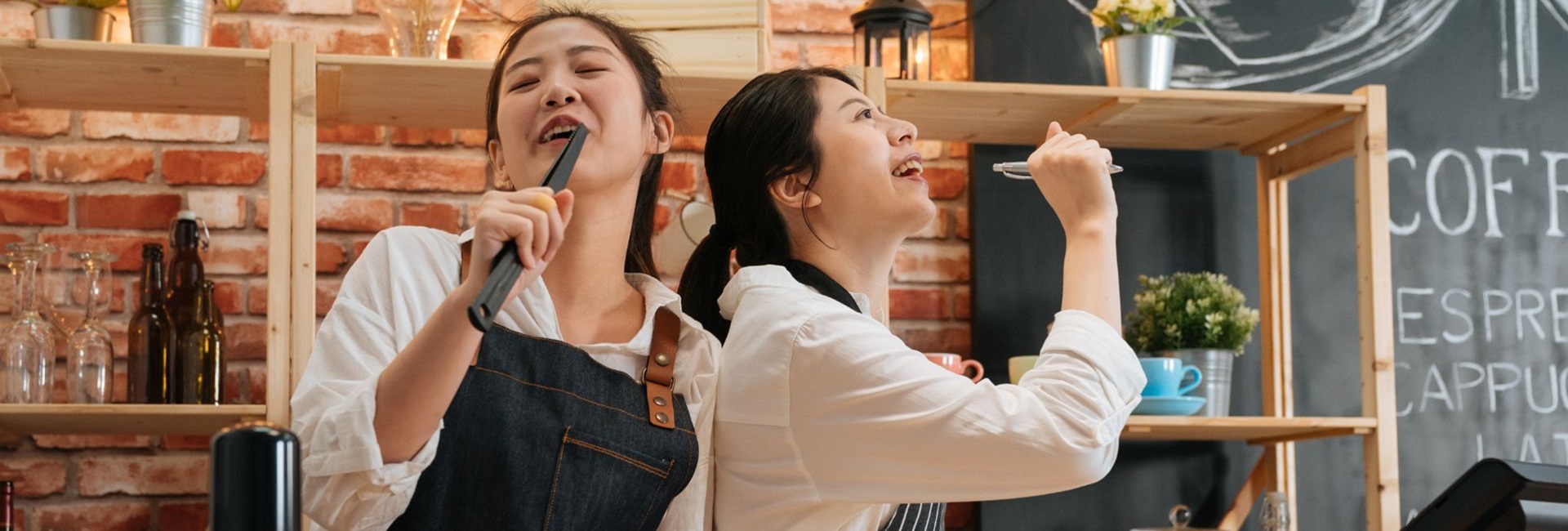 Kaksi nuorta iloista kahvilatyöntekijää palvelutiskin takana esittävät laulavansa. Toisella kädessä salaattiottimet, toisella kynä.