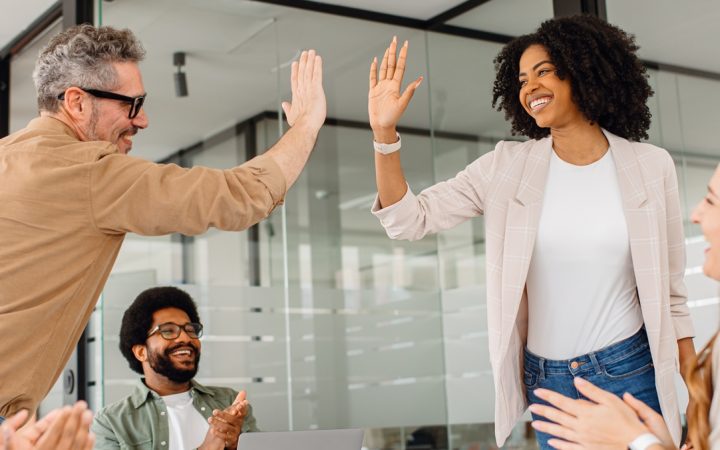 Viisi hyvänntuulista henkilöä työpaikalla pöydän ääressä. Kolme istuu ja taputtaa, kaksi seisoo heittäen toisilleen yläviitosen.