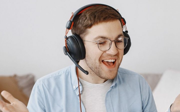 Iloinen mies kuulokkeet päässä opiskelee kannettavalla tietokoneella kotonaan. Vieressä pöydällä myös kirja.