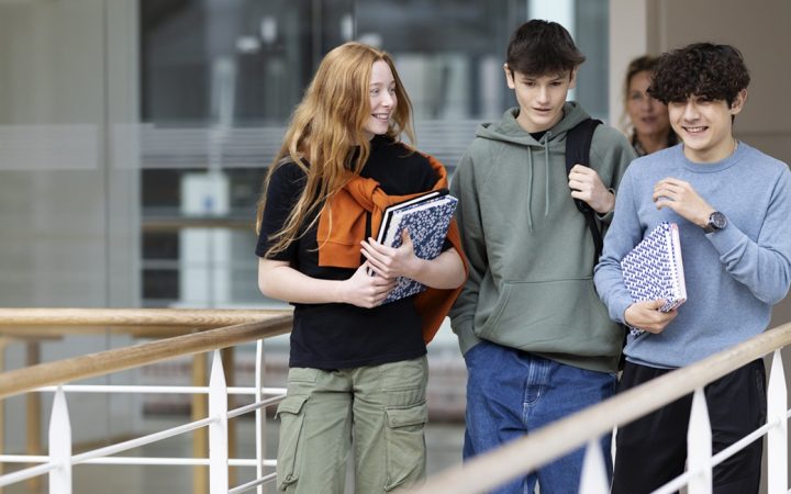 Kolme nuorta opiskelijaa kävelemässä koulun kaiteiden rajaamaa käytävää.