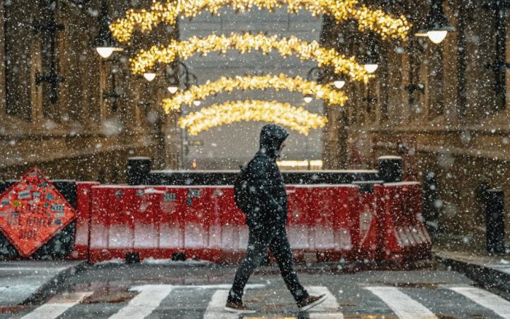 Hupparipäinen nuori ylittää suojatietä harmaassa , räntäsateisessa kaupunkiympäristössä. Taustalla kadun sulkevat betoniesteet. Kadun yli menevät jouluvalot.