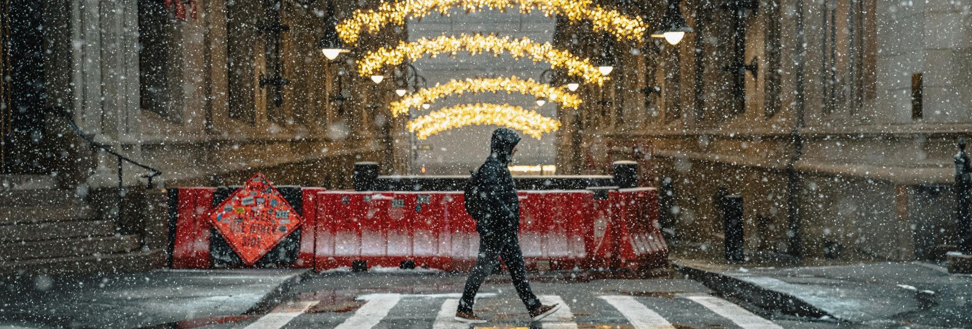 Hupparipäinen nuori ylittää suojatietä harmaassa , räntäsateisessa kaupunkiympäristössä. Taustalla kadun sulkevat betoniesteet. Kadun yli menevät jouluvalot.