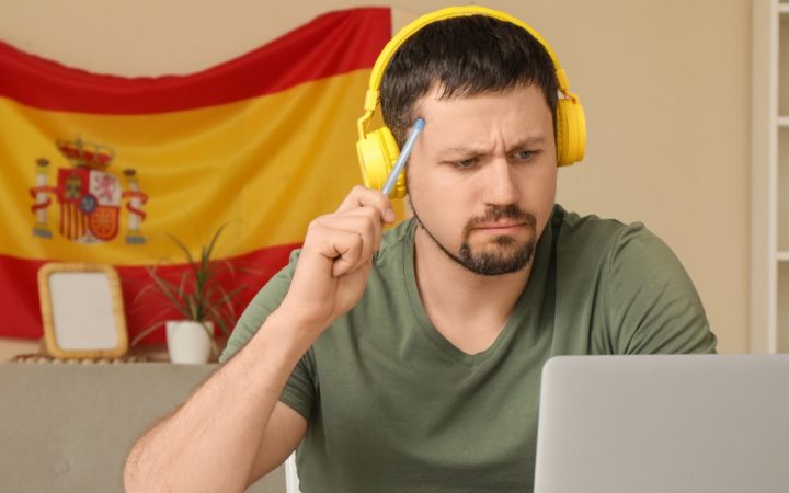 Mies mietteliäänä tietokoneen ääressä opiskelee espanjaa, taustalla Espanjan lippu.