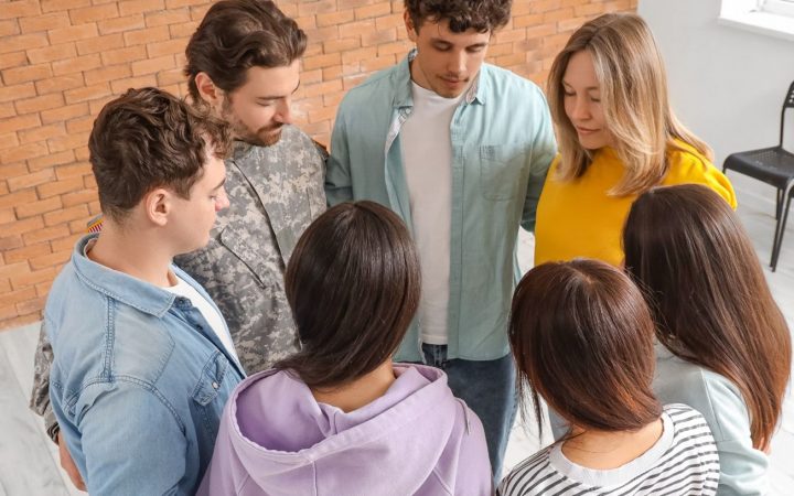 Seitsemän ihmistä seisomassa ringissä silmät kiinni kädet toistensa vyötäröillä.