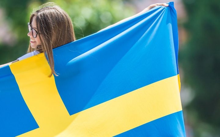 Nainen puistossa kantaa hymyillen Ruotsin lippua ja pitää sitä auki selkänsä takana