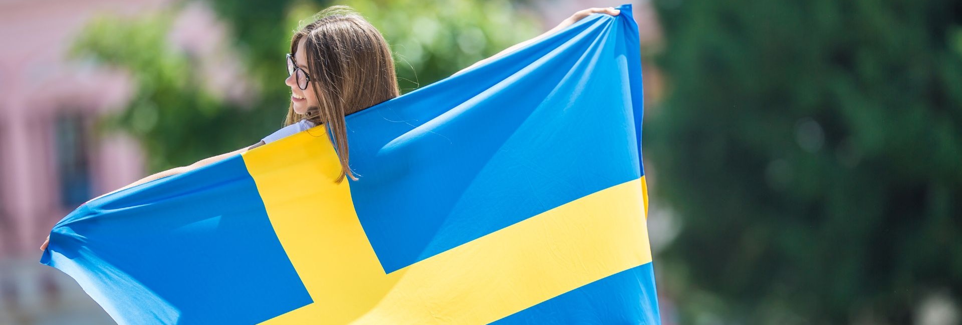 Nainen puistossa kantaa hymyillen Ruotsin lippua ja pitää sitä auki selkänsä takana