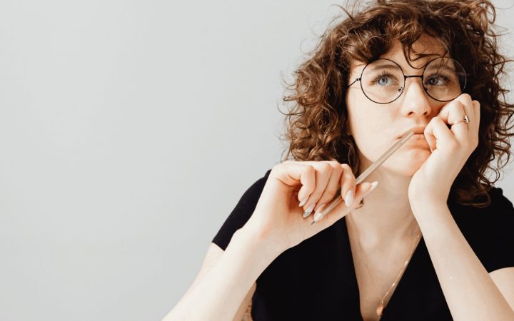 Mietteliäs, silmälasipäinen nainen nojaa toisella kädellään poskeen, toisessa kädessä kynä