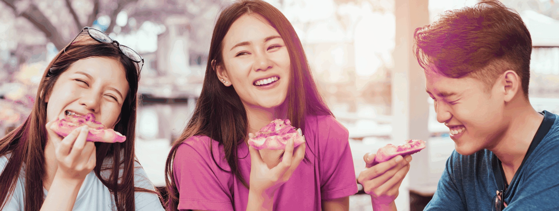 Kolme opiskelijaa istuvat nauraen pöydän ääressä. Jokaisella on kädessään pitsapala.